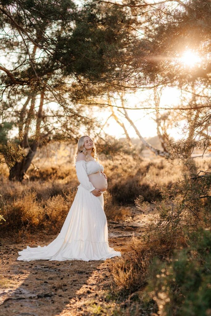 mama im weissen kleid und babybauch stehend lächelt in der lüneburger heide