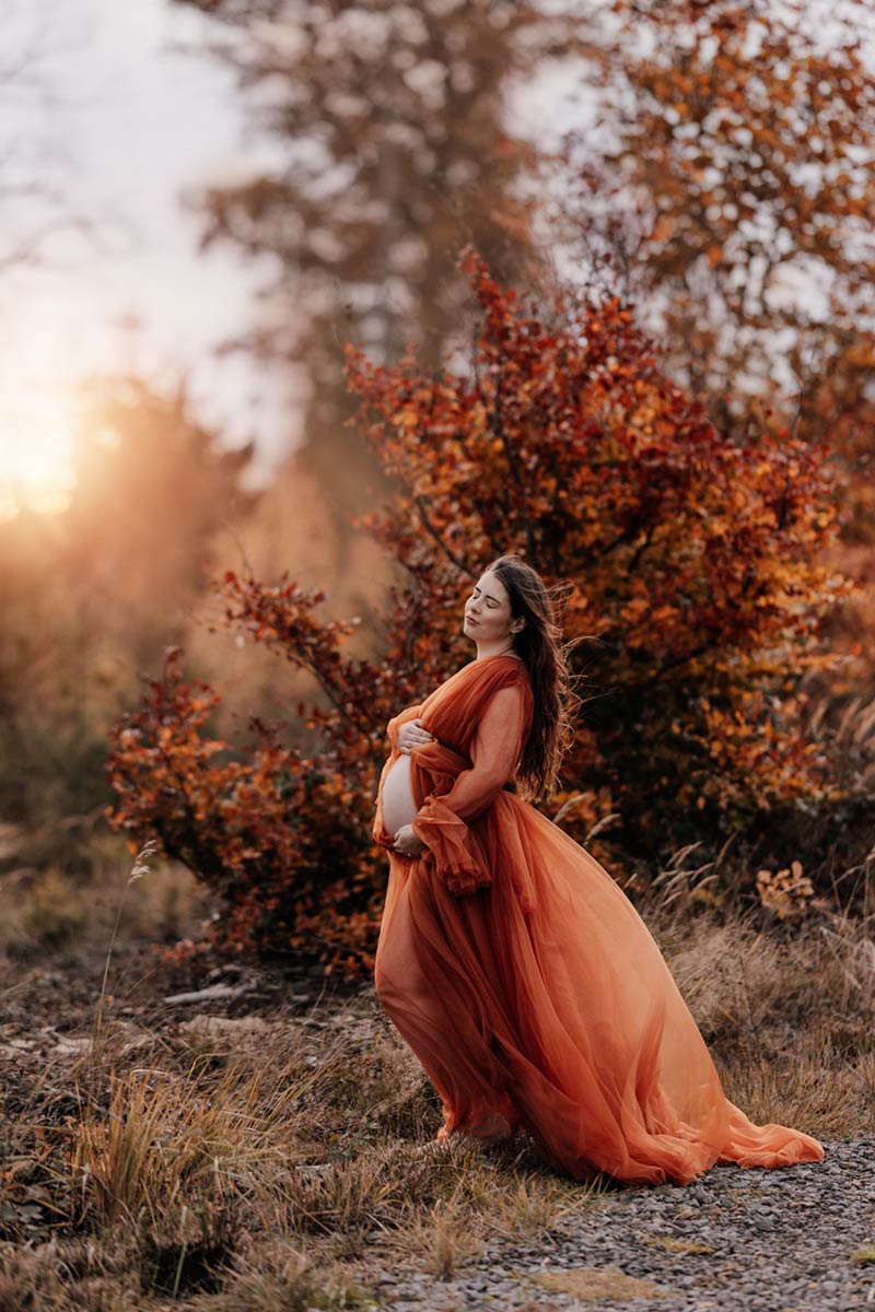 mama mit babybauch steht im orangenen tüllkleid beim fotoshooting zur abendsonne vor eibem herbstlichen busch wald